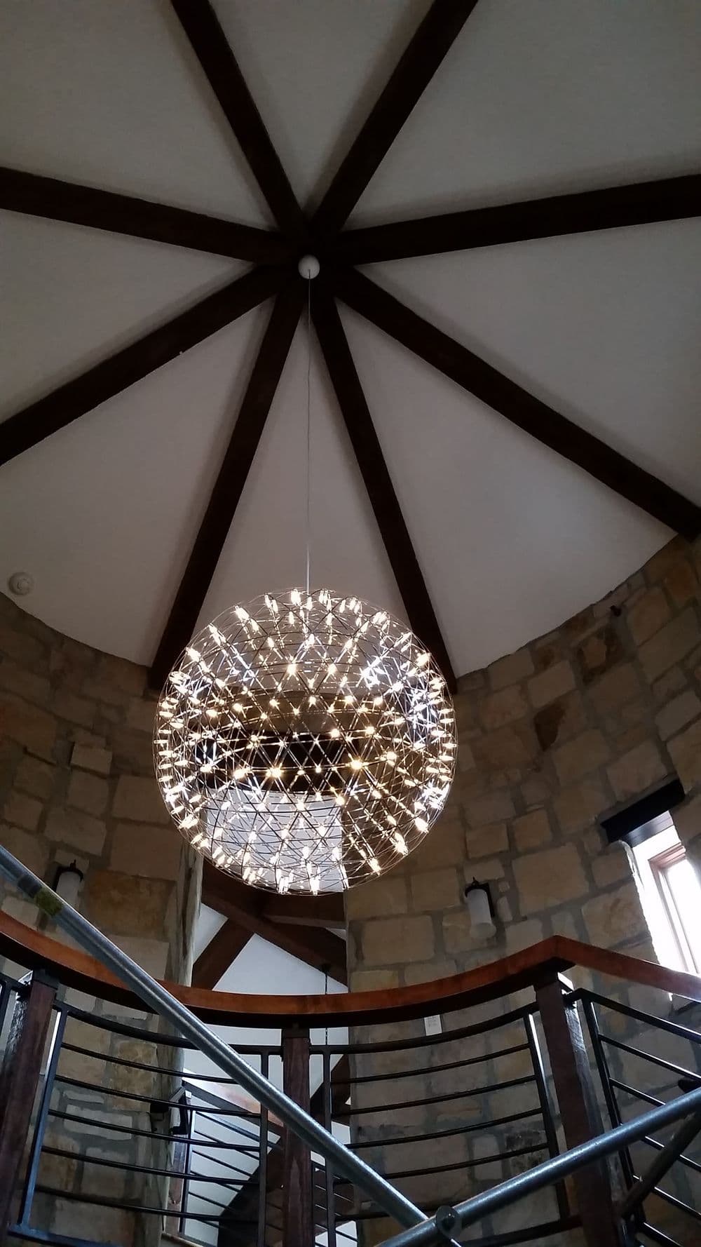 Modern chandelier hanging from a vaulted ceiling with wooden beams in a stone stairwell.