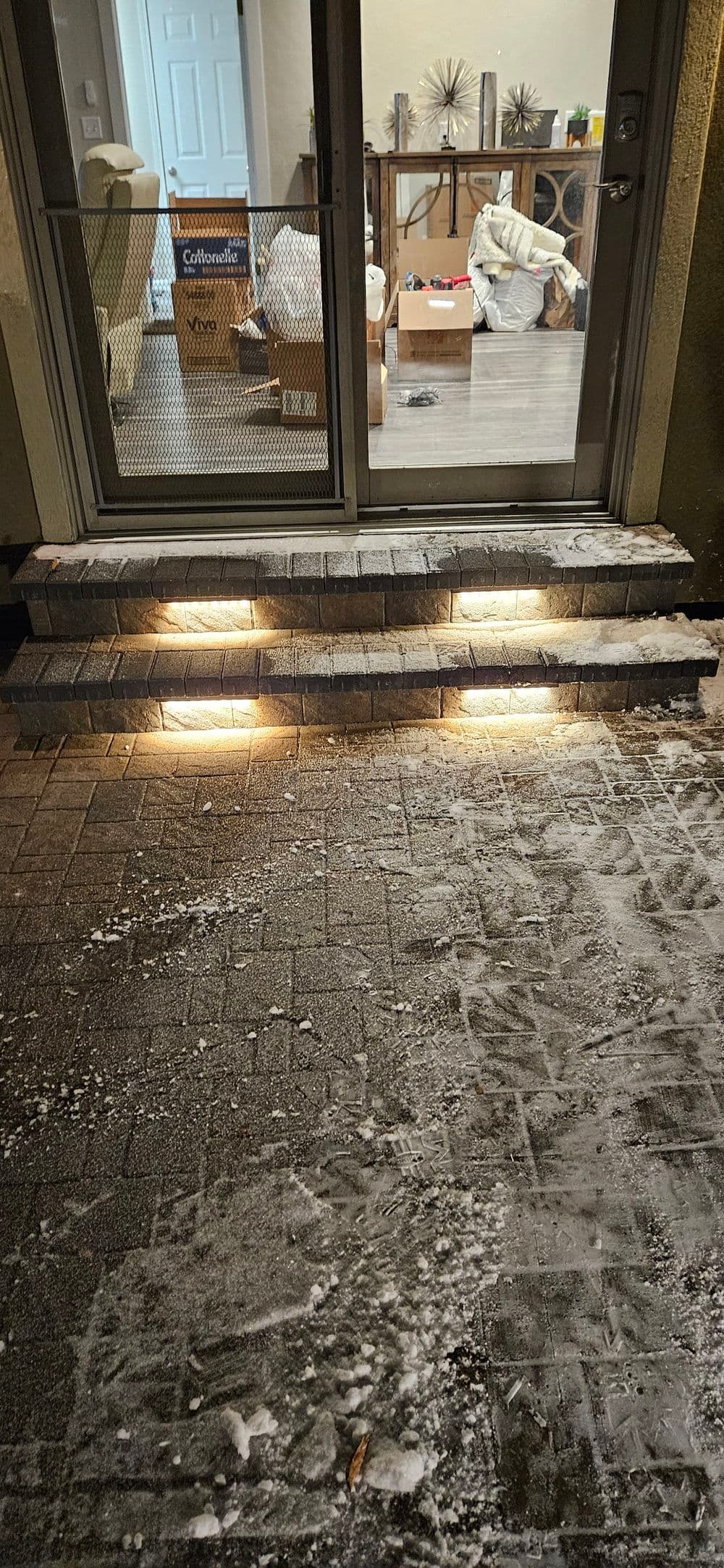Snow-covered steps leading to a doorway with warm lights, indoors visible through the glass.