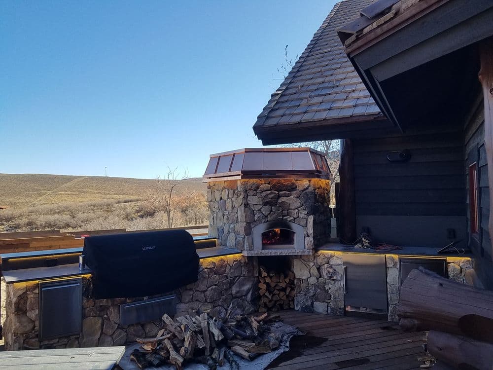 Outdoor stone fireplace with wood storage and grill, set against a clear blue sky.