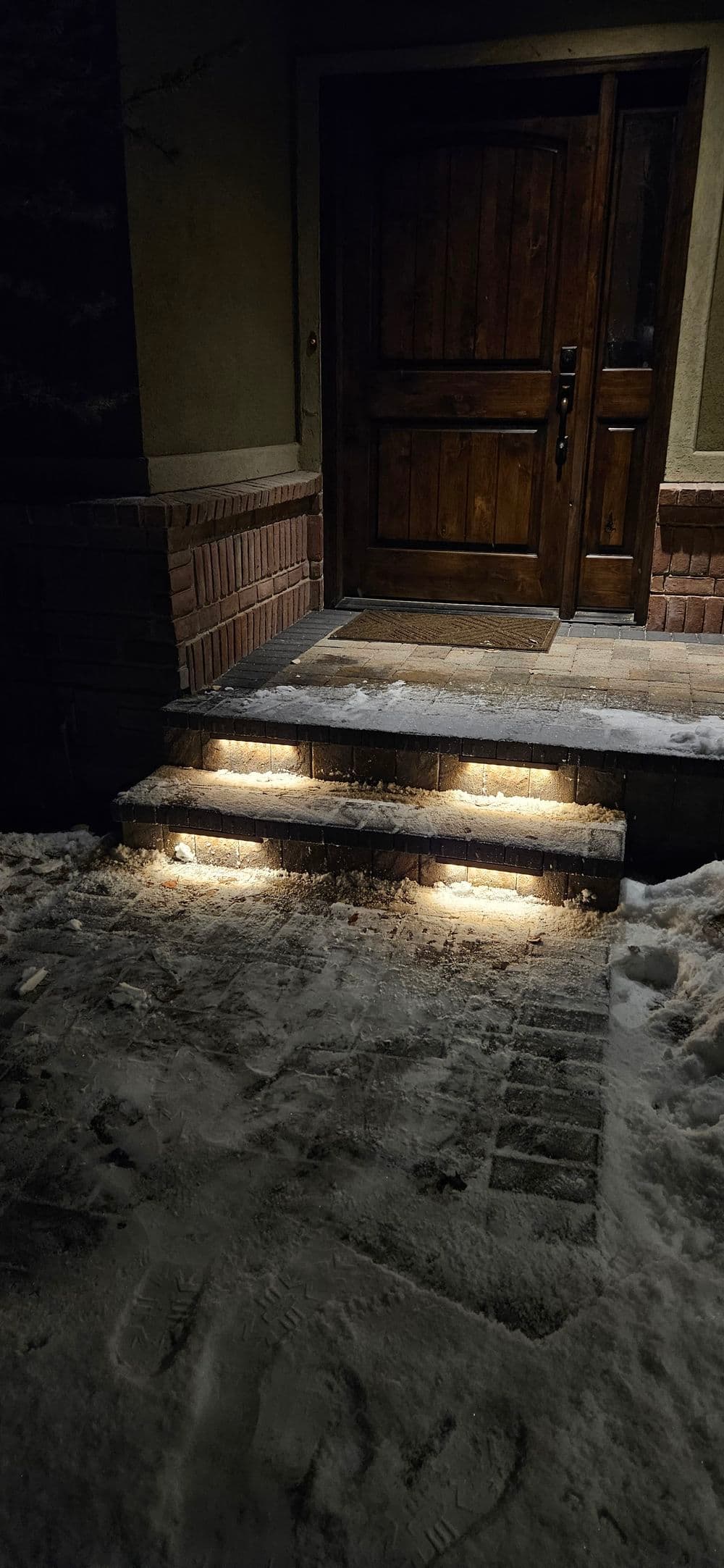 Illuminated entrance steps and wooden door covered in fresh snow at night.