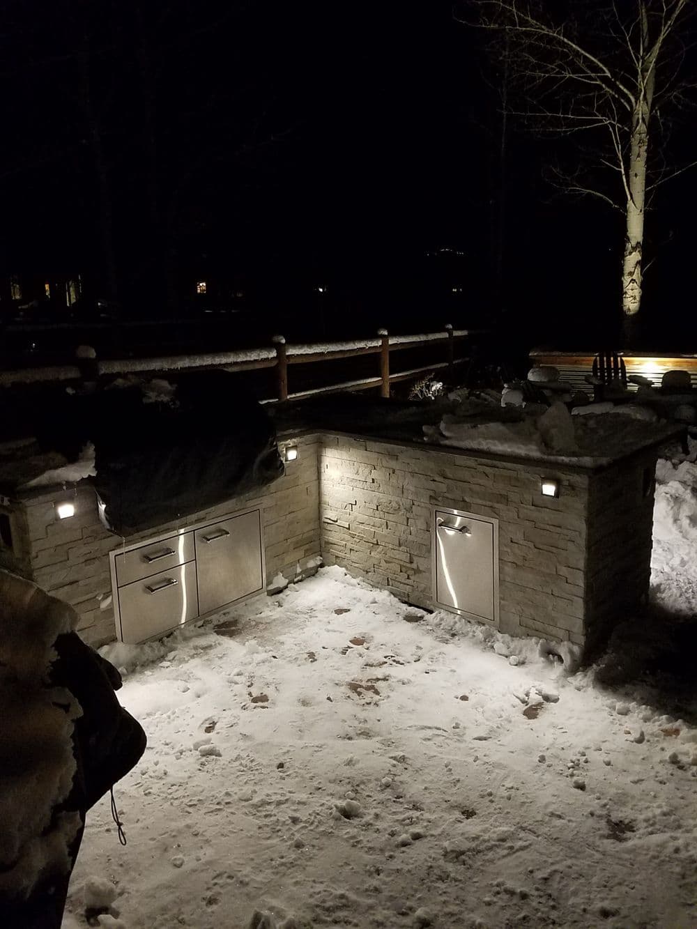 Outdoor kitchen at night, featuring stone construction and illuminated fixtures in snowy setting.