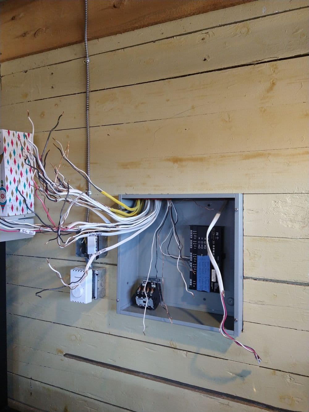 Electrical panel with exposed wires mounted on a wooden wall, showcasing wiring connections.