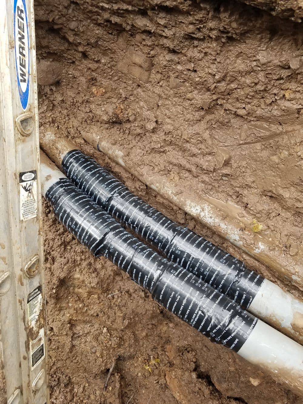 Pipes wrapped with black tape in a muddy trench alongside a ladder.