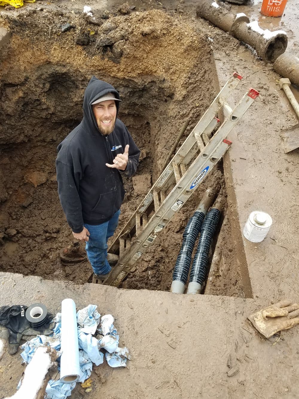 Construction worker smiling near a ladder in a muddy excavation site with pipes.